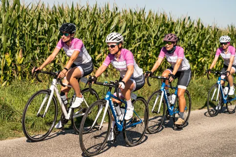 Group of cyclists on a road through a corn field, with t-shirts featuring Bristol Myers Squibb (BMS) and the Country 2 Country 4 Cancer ride