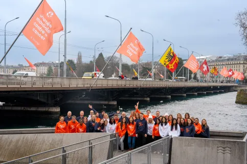 Group of people in World Cancer Day branded white, blue and orange sweatshirts, outside below a bridge flying World Cancer Day flags