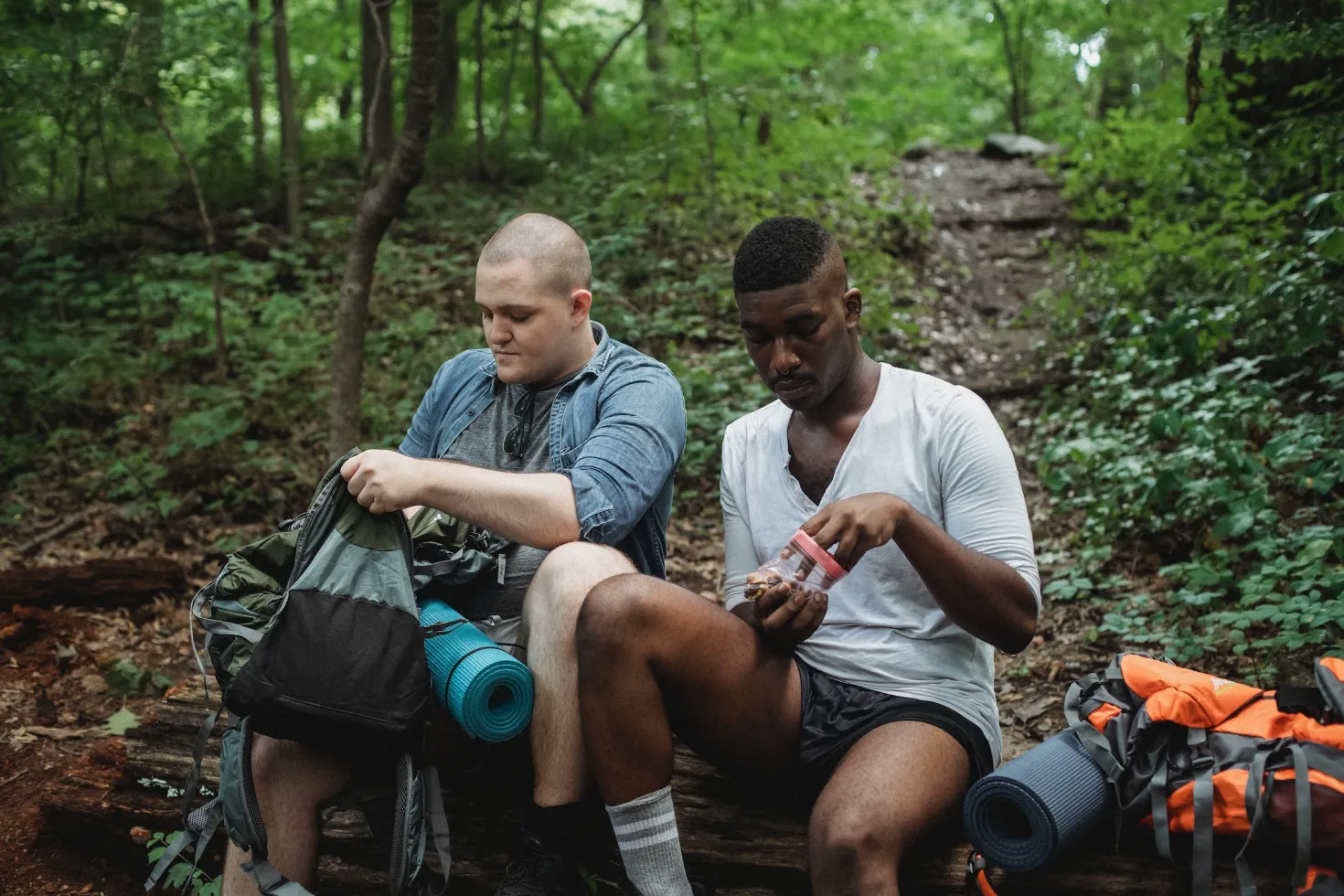 A White and a Black man taking a break to eat a healthy snack while on a hike in a forest