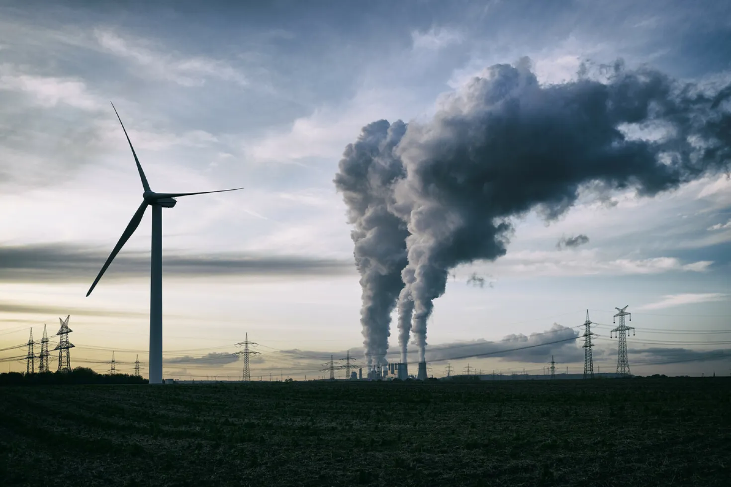 Smoke stacks belching dark smoke next to a wind turbine, at dawn