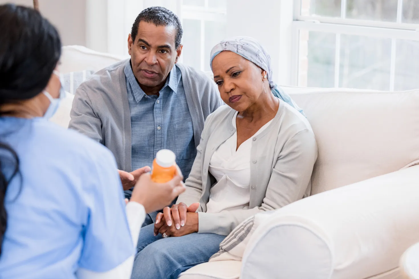 Black middle-aged man and woman holding hands, the woman wearing a headscarf indicating illness, both are listening to a doctor