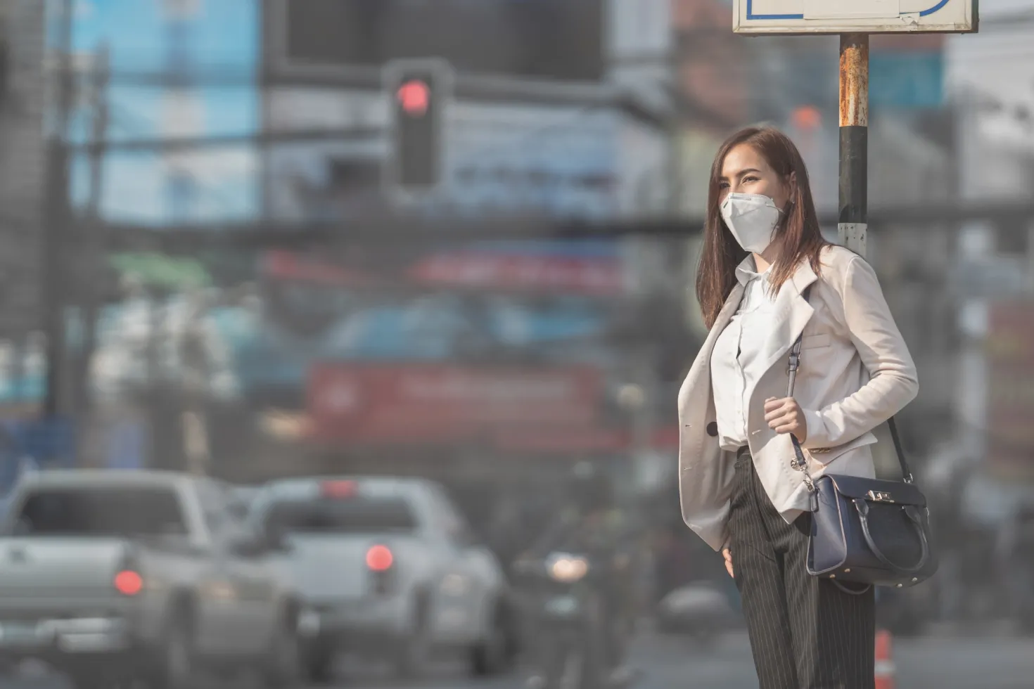 Woman wearing a mask waiting on the sidewalk next to traffic in polluted air