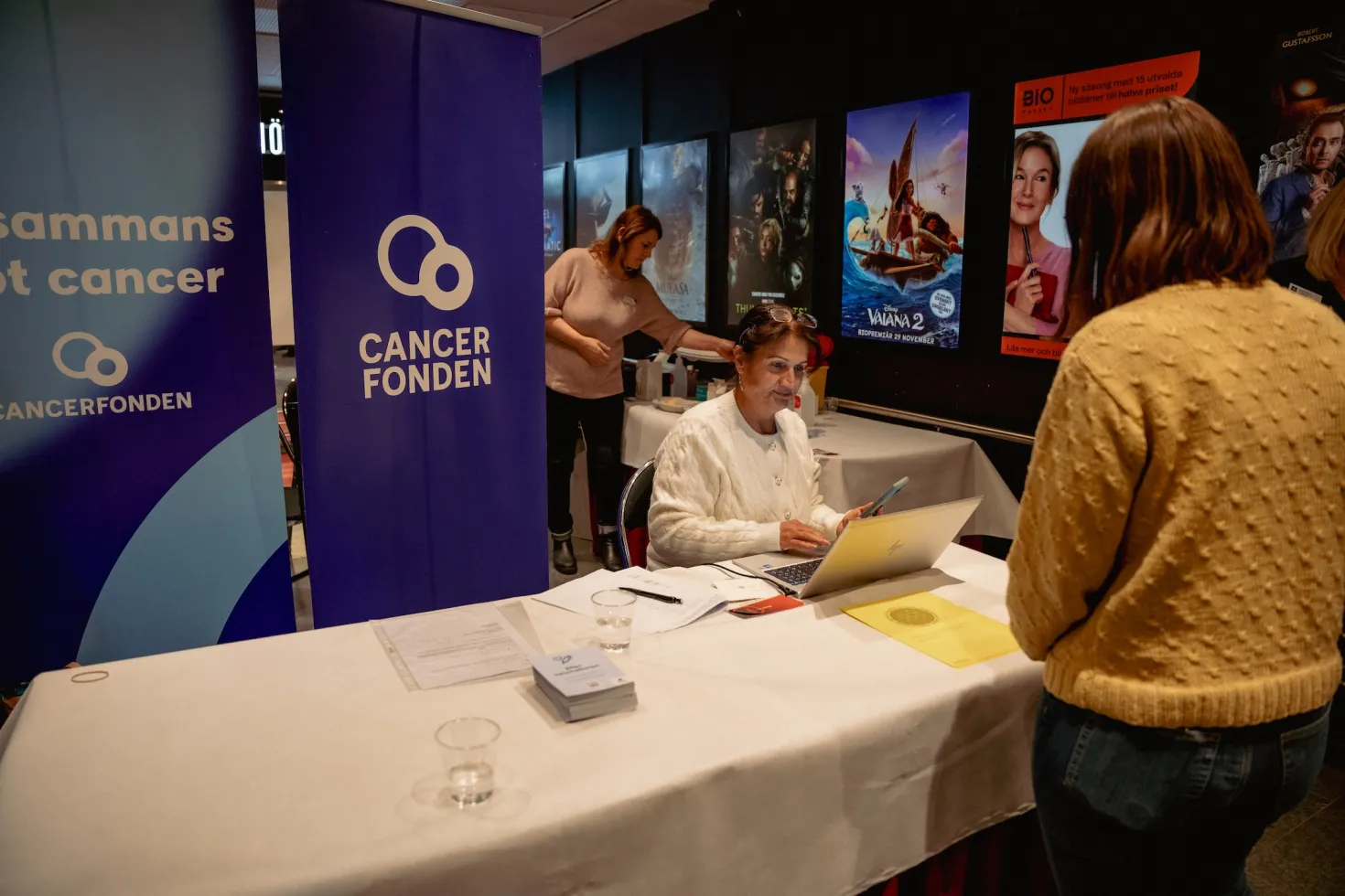Swedish Cancer Society booth at a move theater, a woman sitting down is taking details from a customer