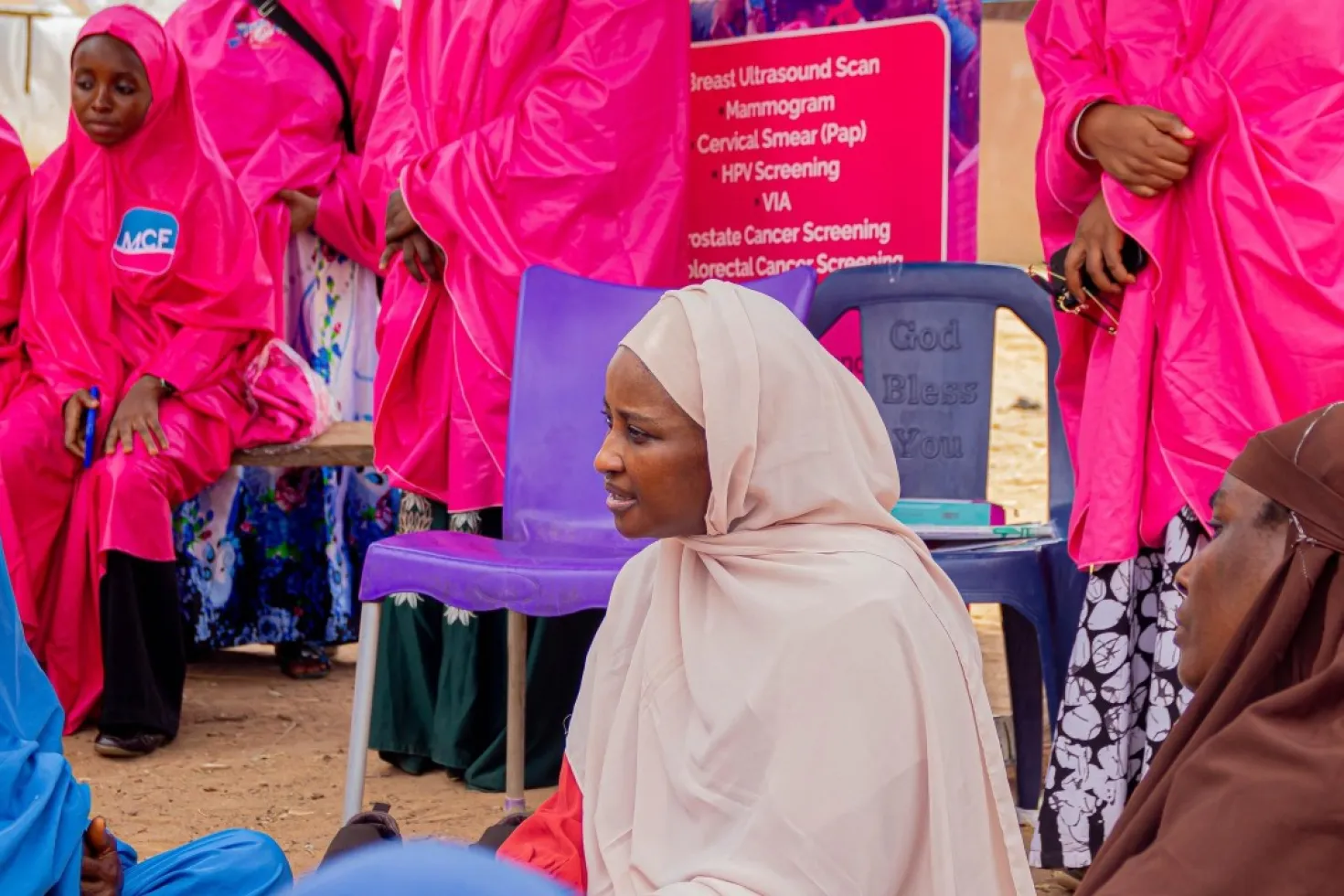 A group of African women wearing bright pink garments, gathered around a sign that lists various medical services, including HPV vaccination for cervical cancer