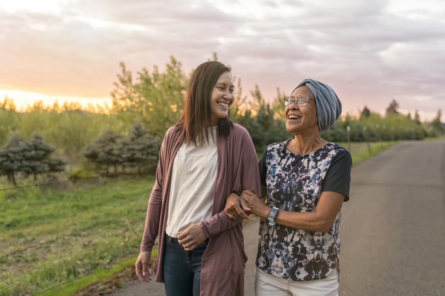 A senior woman with cancer walks with her adult daughter at sunset down a rural road. The affectionate pair are talking and walking with arms linked.