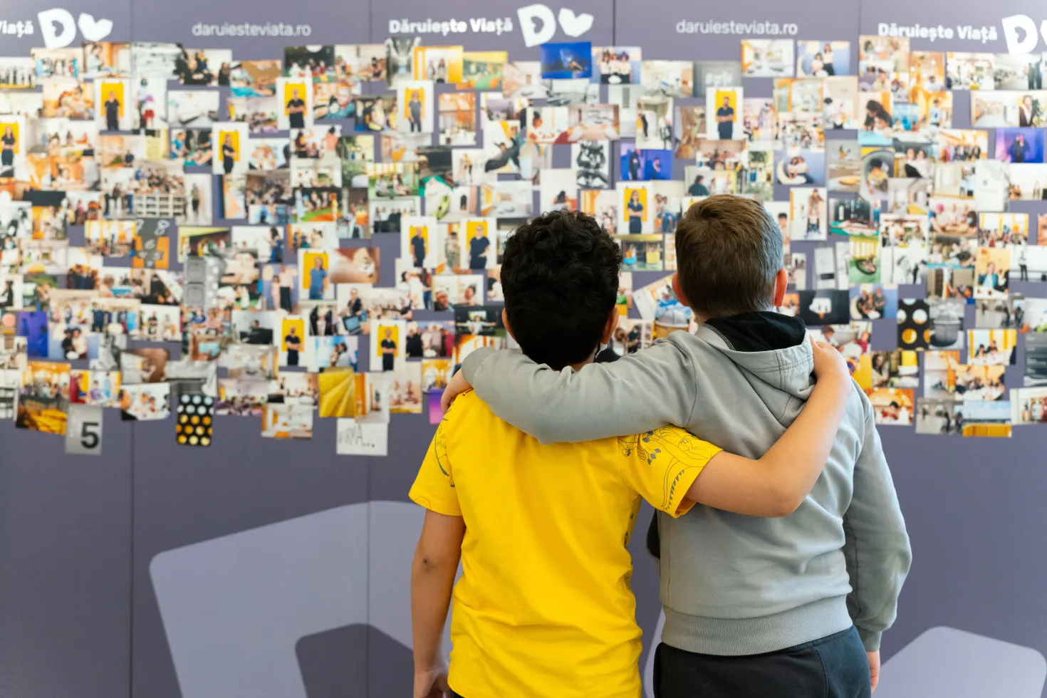 Two children, back to the camera, one in yellow t-shirt, other in great sweatshirt, look at a wall of photos