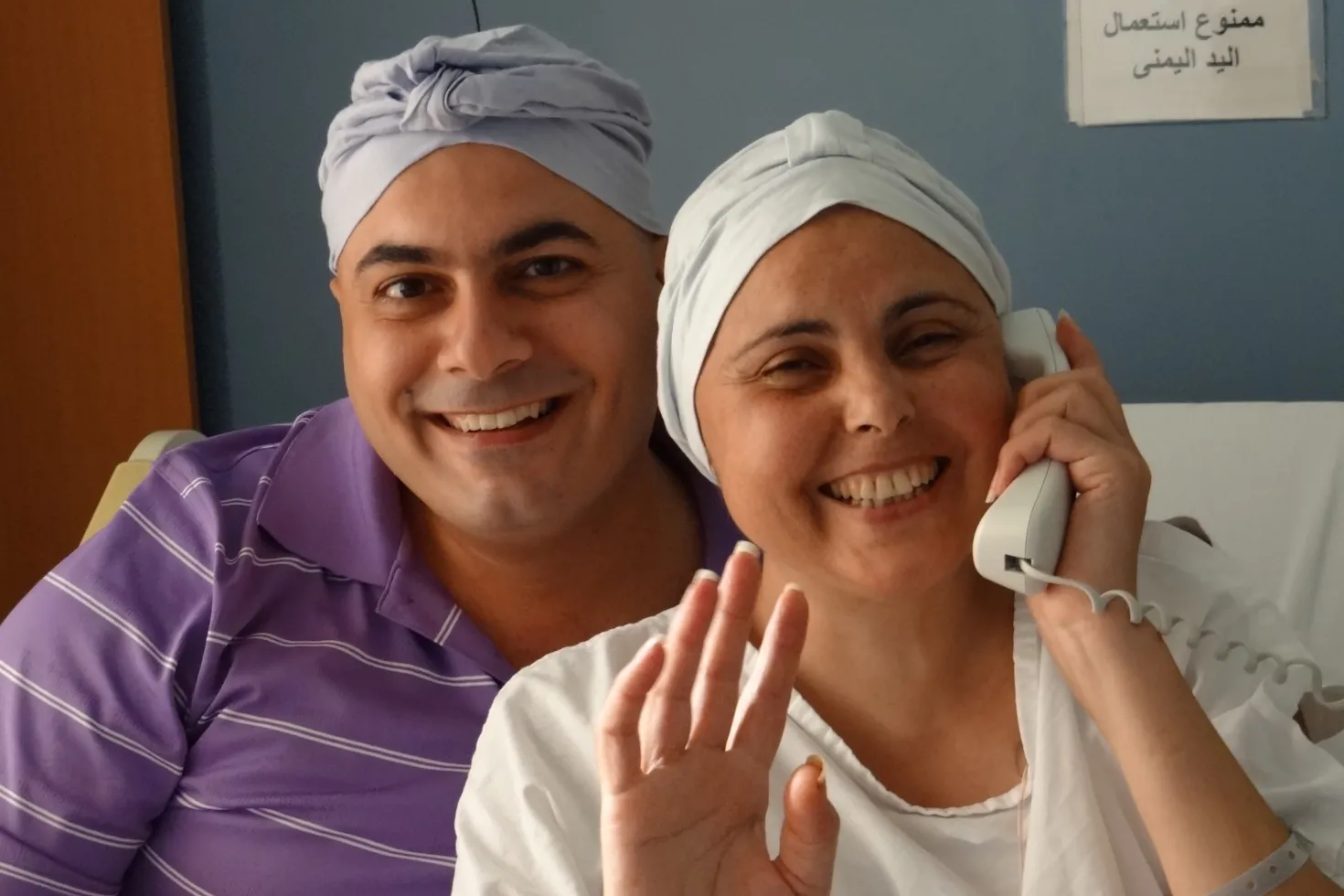 Smiling man wearing a purple t-shirt and headscarf with smiling woman with cancer on the phone, a hand raised, also with a headscarf