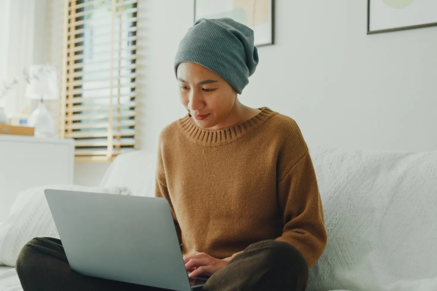 Young Asian woman wearing a headscarf comfortably working on her laptop, seated on a sofa in a well-lit, plant-decorated room.