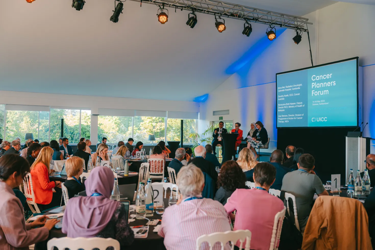 Alt text: A conference setting featuring a panel discussion at the Cancer Planners Forum. Attendees are seated at tables, engaged in the presentation. A large screen displays information about the forum, while speakers are positioned on stage. The room is well-lit with a modern design, and various attendees are seen taking notes or listening attentively.