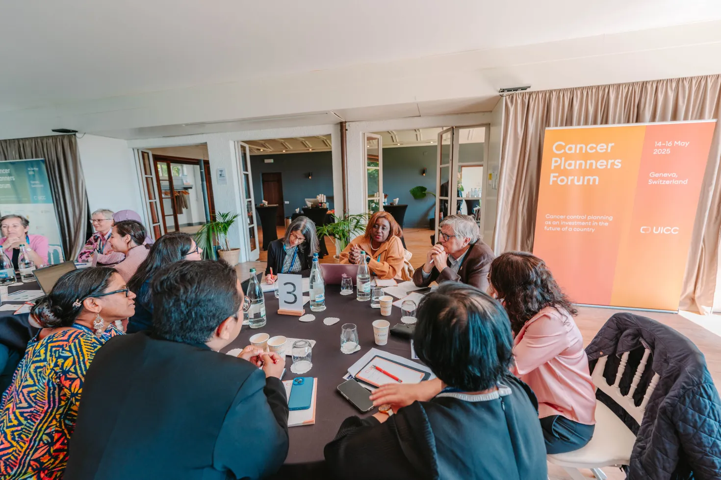 A group of diverse individuals is seated around a round table in a well-lit meeting room, engaged in discussion during the Cancer Planners Forum. The table is adorned with cups, notebooks, and a number card indicating "3." In the background, a large orange banner displays the event's name and details, set against a backdrop of large windows and plants. The atmosphere appears collaborative and focused.