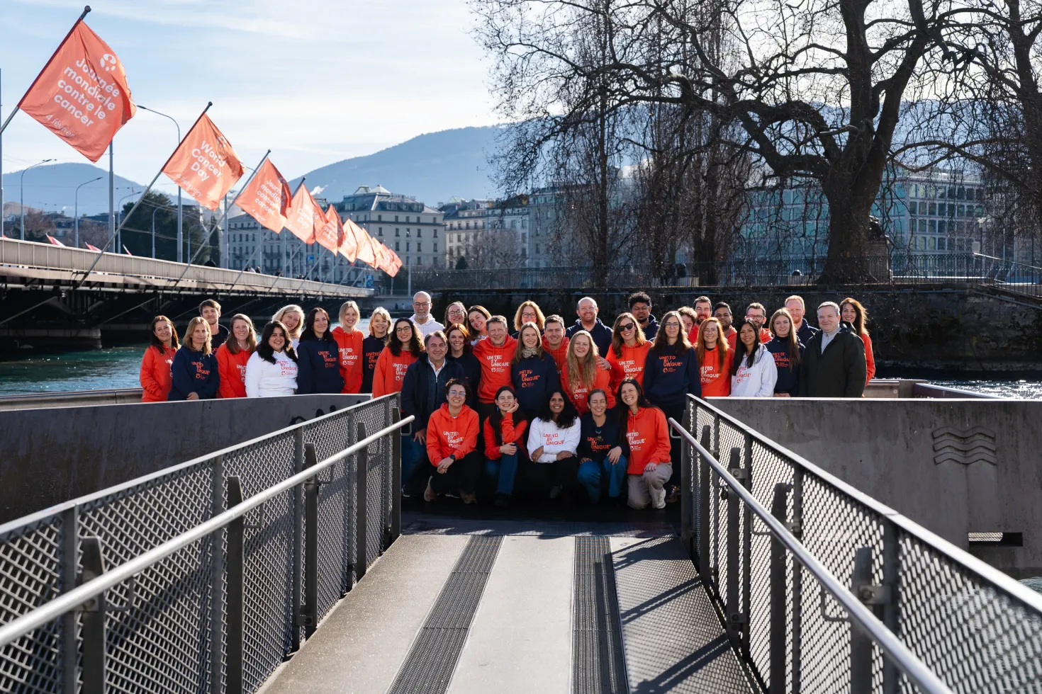 A large group of UICC Staff members, are posing for a photograph on a modern, grey footbridge over a river. They are wearing orange t-shirts with the text "World Cancer Day" printed on them. In the background, a snow-capped mountain range is visible, and several orange flags bearing the "World Cancer Day" logo are displayed along the bridge. The scene is taken at the Mont-Blanc bridge in Geneva, Switzerland