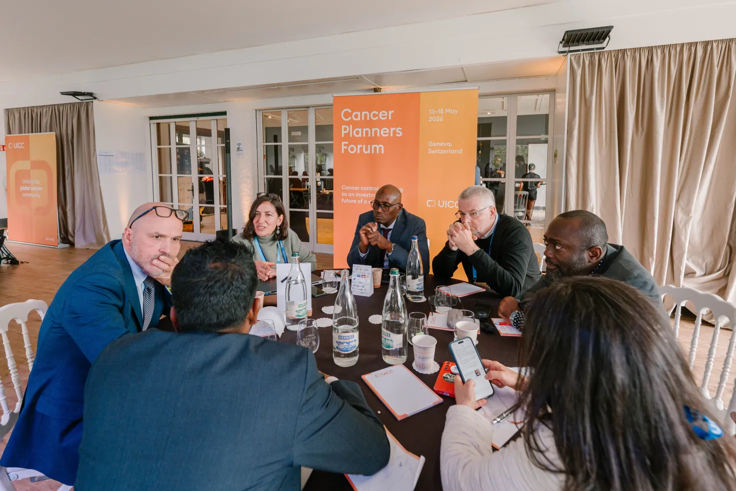 A group of seven professionals are seated around a conference table, engaged in discussion. The table is covered with notepads, pens, and water glasses. A banner behind them reads "Cancer Planners Forum" with dates and location information. The setting appears to be a conference room with a neutral-toned backdrop and a window visible in the background. The UICC logo is present on several materials.