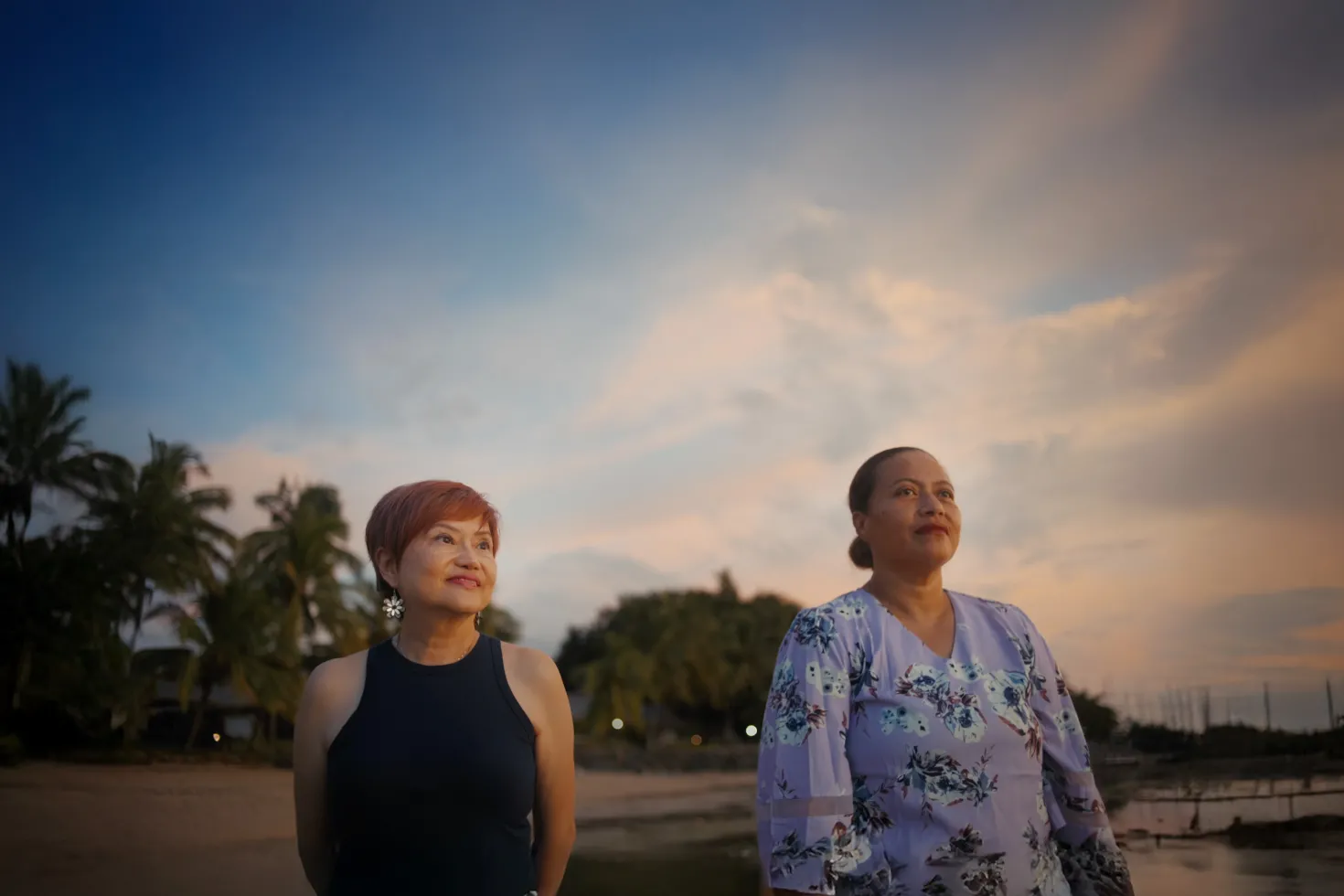 Two people stand side by side outdoors near a shoreline at sunset, looking toward the horizon. The scene is warmly lit with soft golden light and a partly cloudy sky. In the foreground, one person wears a sleeveless dark top and short hair with earrings, while the other wears a light purple floral-patterned dress with hair pulled back. In the background, there are palm trees and what appears to be a marina or moored boats, slightly out of focus, creating a calm coastal setting.
