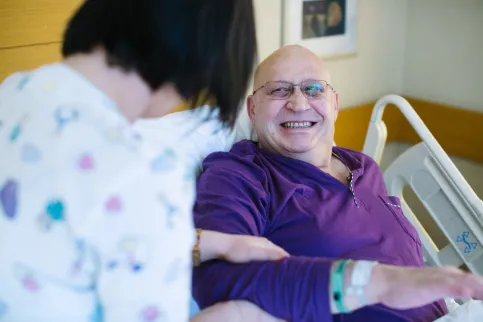 White male elderly patient in purple hospital gown, in a hospital bed in Acibadem Kozyatağı Hospital, smiling while being treated by a female nurse.