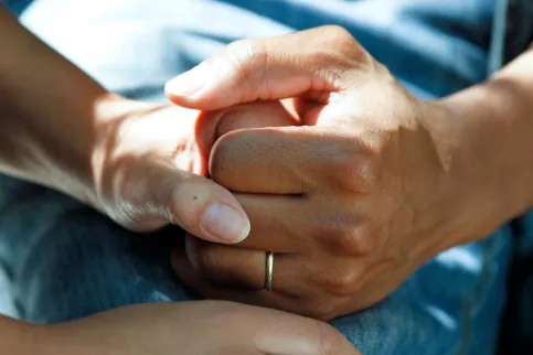 Close up of two hands clasped, resting on a hospital bed, one with a wedding ring