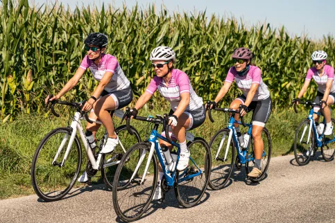 Group of cyclists on a road through a corn field, with t-shirts featuring Bristol Myers Squibb (BMS) and the Country 2 Country 4 Cancer ride