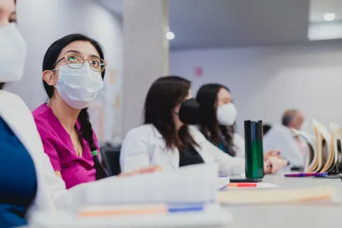 A women in doctor's overall listening to a lecture, sitting at a table with other doctors, all wearing masks