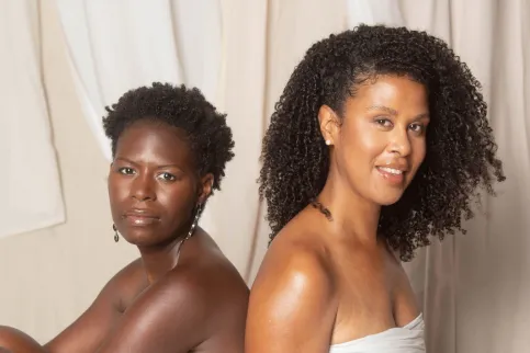 Two Black women back-to-back, cropped just above the breast, heads turned towards the camera