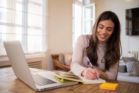 A smiling Hispanic woman writing in a notepad in front of a computer