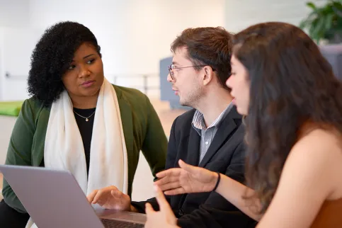A young white man between a young white woman with long hair on his left and a young black woman with a white scarf on his right, talking in front of a computer