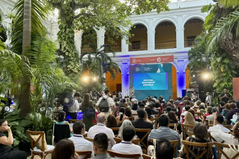 Small crowd outdoors before a white colonnaded building, with palm trees.