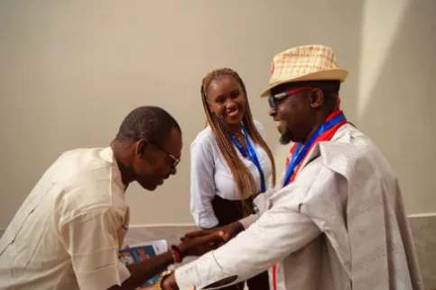 Two Black men and one Black woman, one man is bending before the other, who is wearing a straw hat