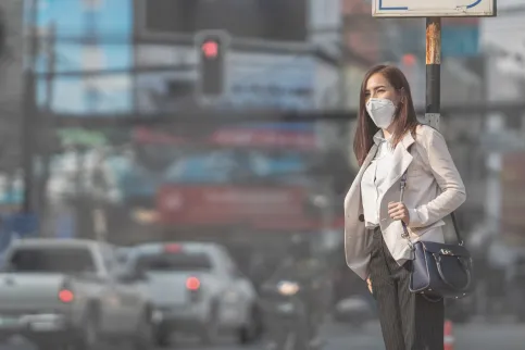 Woman wearing a mask waiting on the sidewalk next to traffic in polluted air