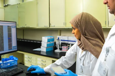 Middle-eastern woman with headscarf and lab coat checking a screen, with a man in lab coat standing just behind her