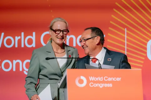 UICC President Ulrika Årehed Kågström wearing a gray suit, smiling with former UICC president Jeff Dunn with a red tie, head turned towards Ulrika, also smiling. both are at the podium at the World Cancer Congress.
