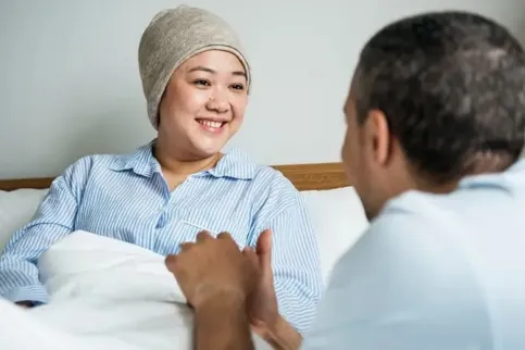 Man seen from behind holding the hand of a smiling woman in a hospital bed, wearing a cap