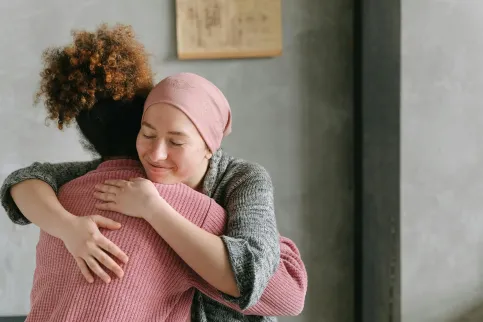 White woman with headscarf hugging a woman wearing a pink sweater