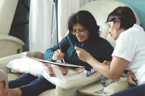 Woman with dark hair and glasses, smiling, is attended by a healthworker as she attempts to draw, while receiving IV treatment and a foot massage