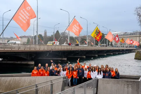 Group of people in World Cancer Day branded white, blue and orange sweatshirts, outside below a bridge flying World Cancer Day flags