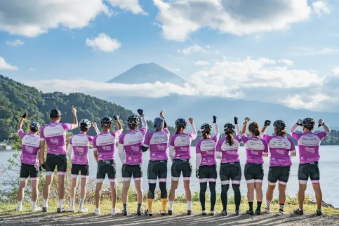 Group of cyclists from Japan in BMS branded pink shirts facing Mount Fuji with arms raised, backs to the camera