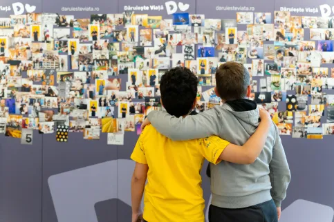 Two children, back to the camera, one in yellow t-shirt, other in great sweatshirt, look at a wall of photos