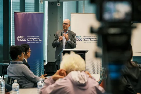 Middle age man wiht no hair, wearing glasses in grey suit with no tie, speaking to a diverse audience
