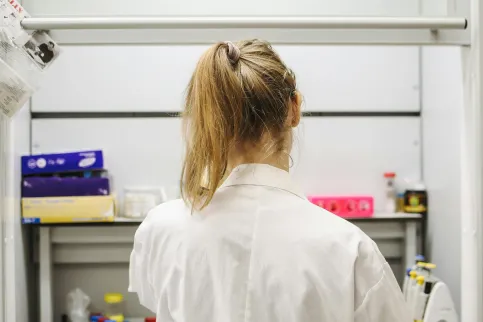Women with long dirty blond hair in a pony tail, in a white lab coat, seen from behind looking at lab equipment