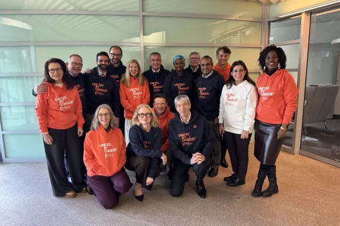 Diverse group of people in two rows, one kneeling in front, posing for the camera in branded white, blue, or orange World Cancer Day United by Unique hoodies