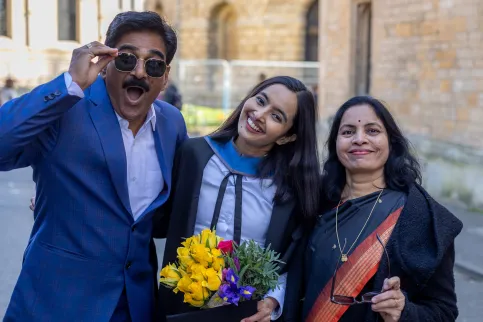 Young Indian woman in graduation gown holding a bouquet of yellow flowers celebrating flanked by her father on her left and mother on her right