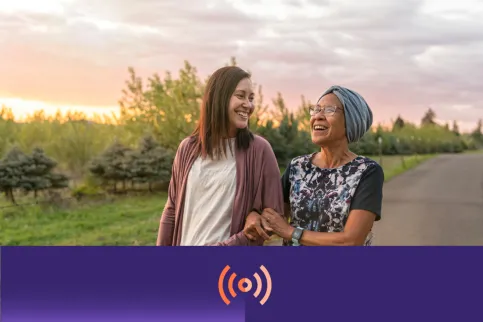 A senior woman with cancer walks with her adult daughter at sunset down a rural road. The affectionate pair are talking and walking with arms linked.