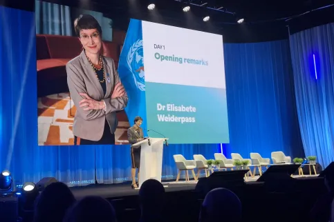 Middle aged white woman at a podium on a big stage with her picture and name Elisabete Weiderpass in the background