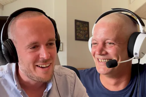 two men wearing headphones with mics, smiling, one without hair, a cancer patient