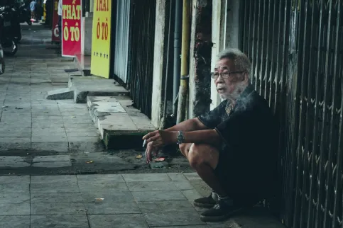 Older man kneeling near a shop, smoking a cigarette