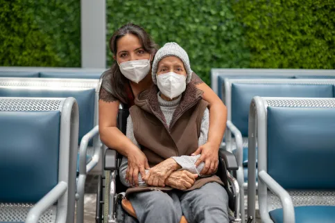 Young woman standing behind and embracing an older woman in a wheelchair, both wearing masks. Photo by Adán Jardón (c) UICC 2021