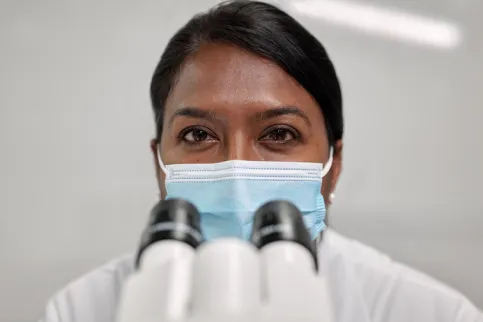 Health practitioner masked looking up from a microscope. 