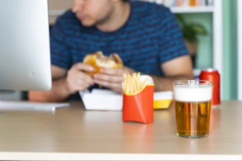 Cropped head of a white male eating burger, fries, soda and beer in front of his computer.