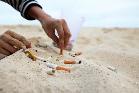 Close up of a person's hand putting out a cigarette in the sand next to other cigarette butts