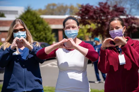 Three women nurses wearing masks and showing a heart sign outside a health facility
