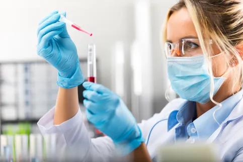 Female scientist in protective eyeglasses, mask and gloves dropping a red liquid substance into the test tube with a pipette in the scientific chemical laboratory