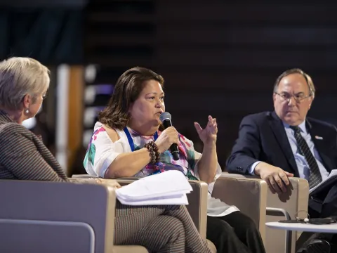 Carmen Auste, CEO of Cancer Warriors Philippines, speaking at a conference panel with two men on either side of her